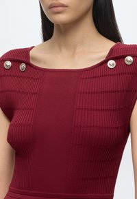 Femme portant un top bordeaux sans manches côtelé avec des boutons dorés décoratifs sur les épaules et un col carré.