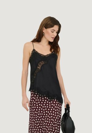 Femme portant un caraco noir orné de dentelle et une jupe marron à pois, tenant un sac à main noir sur un fond uni.