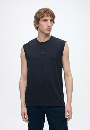 Homme aux cheveux clairs portant un débardeur noir BOSS et un pantalon sombre, debout devant un fond blanc uni, regardant sur le côté.