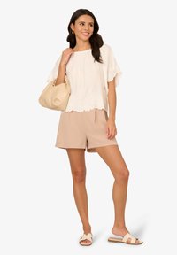 Top bianco testurizzato con orlo ricamato a scallop; pantaloni beige; sandali chiari; borsa beige in mano; stile minimalista, colori tenui.