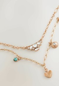 Drei schichtige Halsketten aus Roségold. Die oberste hat klare Edelsteine; die mittlere hat einen Türkis-Akzent; die unterste hat einen Herzanhänger.