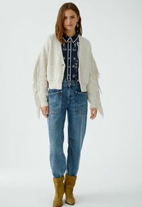 Cremfarbener Strickcardigan mit Fransen, getragen über einem dunkelblauen gemusterten Hemd. Kombiniert mit hellblauen Jeans und gelben Stiefeletten.