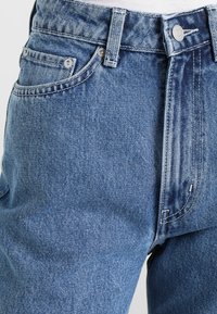 Jeans de ganga azul claro com um design clássico de cinco bolsos, corte reto, costura visível e detalhes em botão prateado na cintura.