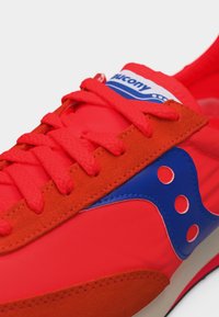 Φωτεινό κόκκινο παπούτσι Saucony με μπλε λογότυπο, κόκκινα κορδόνια και πορτοκαλί σουέτ λεπτομέρειες.