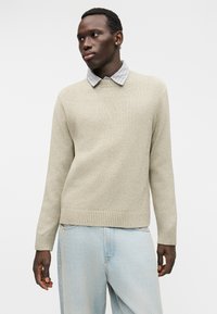 Μπεζ πουλόβερ πλεκτό με υφή, με ριβέ τελείωμα και μανσέτες, φορεμένο πάνω από ένα ελαφρύ πουκάμισο με καρό γιακά. Παντελόνι ανοιχτό μπλε τζιν.