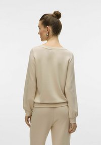 Top beige a maniche lunghe realizzato in morbido tessuto con una vestibilità rilassata, caratterizzato da un ampio scollo e polsini arricciati, abbinato a pantaloni coordinati.
