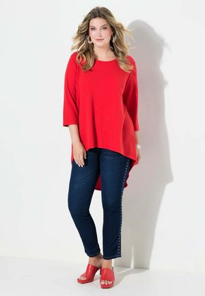 Rotes, langärmliges Tunika-Top mit Rundhalsausschnitt. Slim-Fit, dunkle Jeans mit seitlichen Nieten. Rote, offene Clogs mit einem klobigen Absatz.
