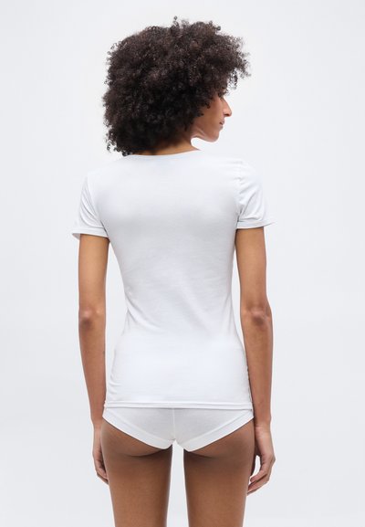 Γυναίκα με σγουρά μαλλιά, φορώντας λευκό απλό κοντομάνικο μπλουζάκι και λευκά εσώρουχα, στέκεται γυρισμένη με την πλάτη προς τα εμάς, μπροστά σε λευκό φόντο.