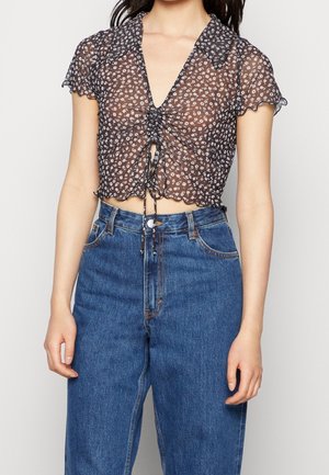 Femme portant un chemisier court noir transparent à fleurs avec manches courtes et jean bleu taille haute sur fond uni.