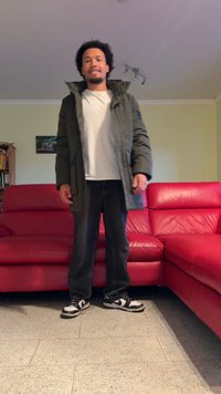 Doudoune verte, t-shirt blanc, jeans noirs et baskets noir et blanc. Debout dans une pièce avec un canapé en cuir rouge.