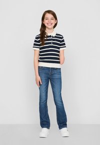 Polo tricot rayé bleu marine et blanc à manches courtes, associé à un denim bleu et des baskets blanches. L'enfant affiche un sourire amical.