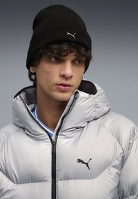 Silver puffer jacket with a high collar and black zipper, features a black Puma logo on the chest. Black knit beanie worn atop curly hair.