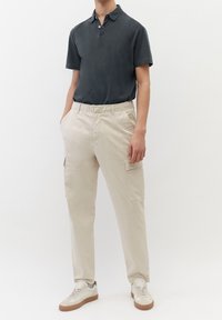 Polo shirt gris foncé avec col et deux boutons, associé à un pantalon cargo crème avec poches latérales. Les chaussures sont blanches avec des accents beiges.