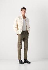 Lichtbeige bomberjack met een hoge kraag, gecombineerd met olijfgroene broek. Het model draagt een witte geribbelde tanktop en zwarte instappers.