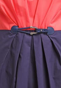 Robe color-block avec un haut corail et un bas marine, dotée d'une texture plissée et d'une ceinture en cuir marine ornée d'une boucle dorée.