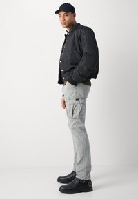 Black bomber jacket, grey cargo pants, black ankle boots with buckle, and a navy cap. Smooth fabric, utilitarian design with pockets.