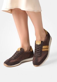 Baskets en suède marron avec des accents beiges, tissu texturé, lacets contrastants et semelle en caoutchouc. Présente un logo sur la languette et une bande sur le côté.