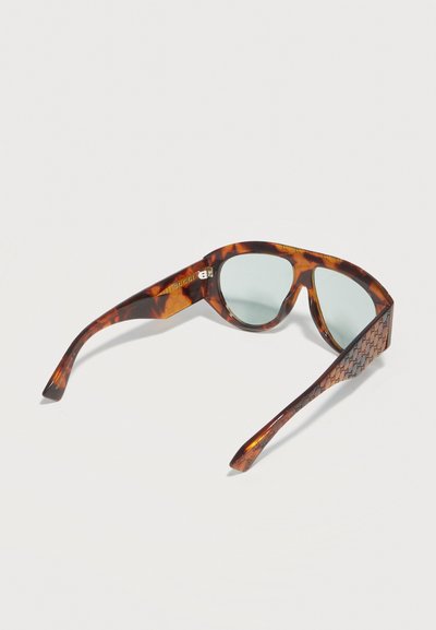 Lunettes de soleil œil de chat en écaille de tortue avec verres bleu clair, logo Gucci sur les branches, et motif G entrelacé sur les branches.