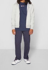 Grauer Reißverschluss-Hoodie über einem dunkelblauen T-Shirt, kombiniert mit dunkelblauen Hosen und weißen Sneakers mit roten und blauen Akzenten. Einfaches Design, lässige Passform.