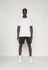 Hvit bomull T-skjorte kombinert med svarte shorts. Modellen har på seg hvite joggesko, og viser rene linjer og et enkelt design. Ingen synlige mønstre eller aksenter.