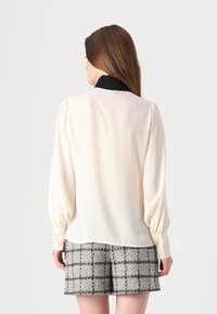 Kremfarget bluse med svart krage, med puffermer og blonder på mansjettene. Kombinert med grå og svart rutete tweed-shorts.