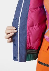 Denim bomberjack met een roze gevoerde voering, witte rits, blauwe drukknoopjes en geribde groene manchetten. Hand die de rand van de jas vasthoudt.