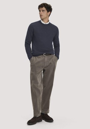 Junger Mann steht mit den Händen in den Taschen, trägt einen marineblauen Pullover, ein weißes Hemd mit Kragen, braune Cordhosen und dunkelbraune Schuhe.