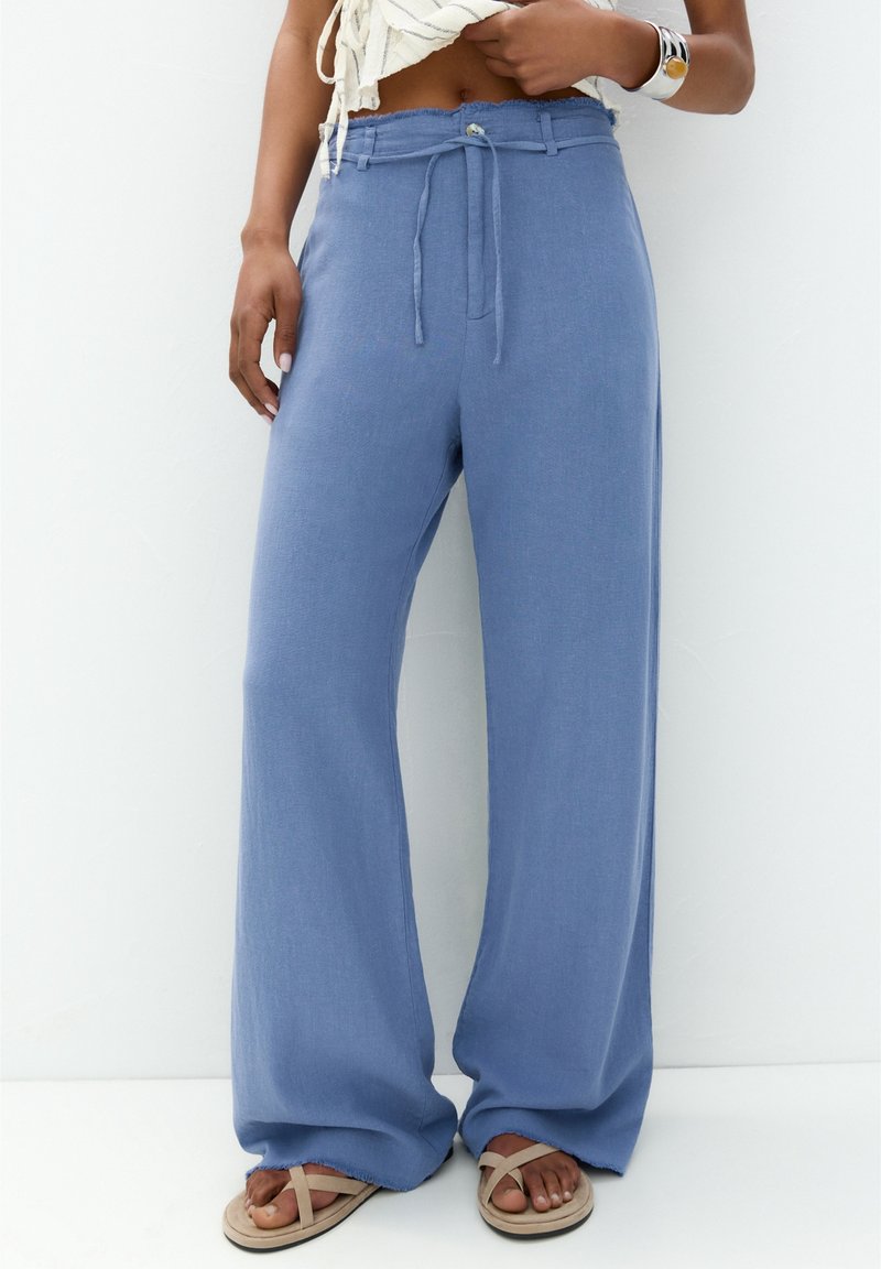 Femme portant un pantalon bleu ample avec une taille à cordon et des sandales beiges, debout devant un fond blanc uni.