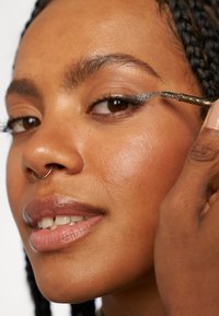 Närbild på en person som applicerar silver eyeliner. Funktionen har slät hud, en septumring och en naturlig läppfärg. Bakgrunden är helt vit.