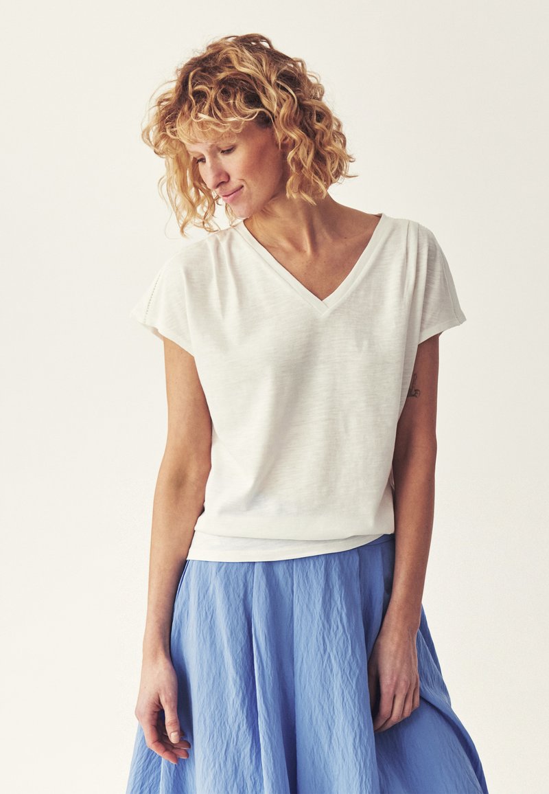 Femme aux cheveux blonds bouclés portant un haut blanc à manches courtes en V et une jupe bleue texturée, regardant vers le bas et souriant doucement.