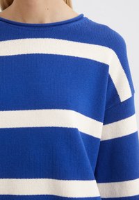 Blauer Strickpullover mit horizontalen weißen Streifen, Rundhalskragen und lockerer Passform. Die Textur wirkt weich mit einem detaillierten Strickmuster.