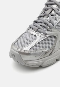 Jasnoszary but sportowy z siateczkową cholewką, metalicznymi akcentami i teksturowaną podeszwą. Zawiera szare sznurowadła i perforacje wentylacyjne.