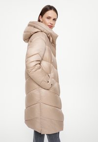 Beige pufferjas met een capuchon, voorzien van een gestikt ontwerp, zijzakken en een gladde, glanzende textuur.