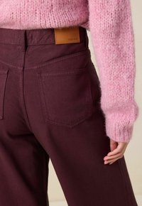 Pantalons larges bordeaux avec deux poches arrière, patch en cuir étiqueté "SISSY-BOY", associés à un pull tricoté rose texturé.