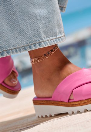 Pink slide sandal with a crossed strap design, wooden-like platform, and white rubber sole. Worn with light denim pants and anklet.