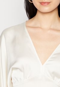 Krämfärgad silkestopp med V-ringning, korta ärmar och rynkdetaljer vid midjan, med en slät och glänsande yta.