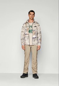 Fleecejakke med et flerfarget abstrakt mønster, beige t-skjorte med grafisk trykk, khaki bukser, og svart fottøy, stående mot en ensfarget bakgrunn.