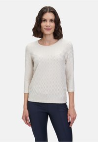 Haut à manches longues texturé, de couleur beige clair, avec un décolleté en forme de scoop et un motif géométrique. Porté avec un pantalon foncé et ajusté. Tissu lisse avec un léger éclat.