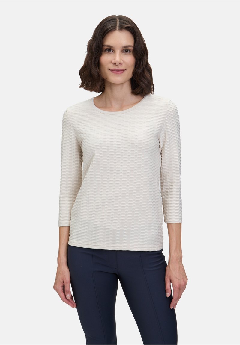 Haut à manches longues texturé, de couleur beige clair, avec un décolleté en forme de scoop et un motif géométrique. Porté avec un pantalon foncé et ajusté. Tissu lisse avec un léger éclat.