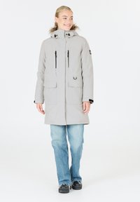 Hellgrauer Parka mit einem mit Fell gefütterten Kapuzen, durchgehendem Reißverschluss, zwei großen Vordertaschen und schwarzen Reißverschlusselementen. Trägt ihn mit ausgebleichten blauen Jeans.