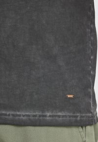 Dunkelgraues Baumwoll-T-Shirt mit leicht strukturierten Oberfläche, ausgestattet mit einem kleinen bronzeartigen Etikett am Saum und einem geraden Schnitt am unteren Rand.