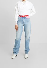 Person som bär ljusblå högmidjade jeans med rosa detaljer och röd bälte, vit zip-up tröja och vita klackstövlar, stående inomhus.