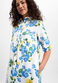 Blumenhemdkleid aus leichtem Stoff; mit blauen Blumen und gelben Zitronen auf weißem Hintergrund; kurze Ärmel, Knopfleiste, Taschen auf der Vorderseite.