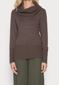 Brązowy sweter dziewiarski z golfem i prążkowaną fakturą, w połączeniu z luźnymi oliwkowymi spodniami. Materiał wydaje się miękki i ciepły.