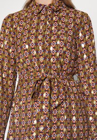 Långärmad klänning med hög krage, med ett flerfärgat geometriskt mönster i rött, blått, gult och guldtoner, med en cinchad midjedetalj.