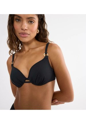 Kobieta o kręconych włosach, ubrana w czarny top bikini z złotymi ozdobami w formie pierścieni, pozuje z jedną ręką za plecami na jasnym, jednolitym tle.