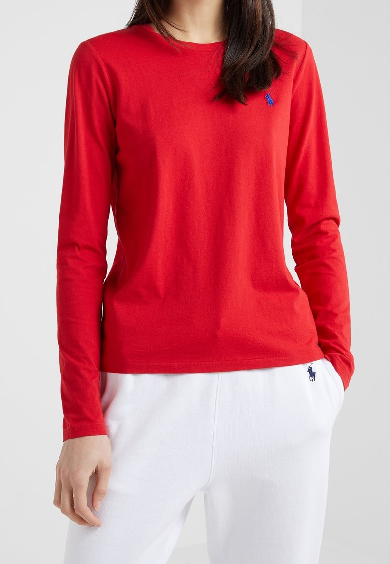 T-shirt rouge à manches longues en coton doux, avec un col rond et un petit logo bleu sur la poitrine, associé à un pantalon de jogging blanc.