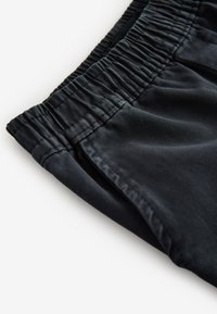 Čierne látkové šortky s textúrovaným elastickým pásom, hladkým povrchom a detailom bočného vrecka, mierne zložené do uhla pre vystavenie.