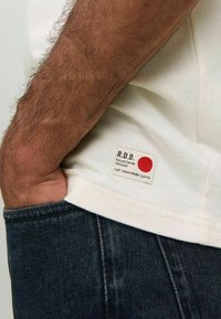 Krämfärgad bomulls-t-shirt med en rektangulär etikett som har en röd punkt och text, i kombination med mörka denimjeans. Fokusera på etikettdetaljer.