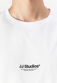 T-shirt blanc à manches courtes avec un col rond, fabriqué en coton doux, avec le logo "JJ Studios" brodé en noir sur la poitrine.